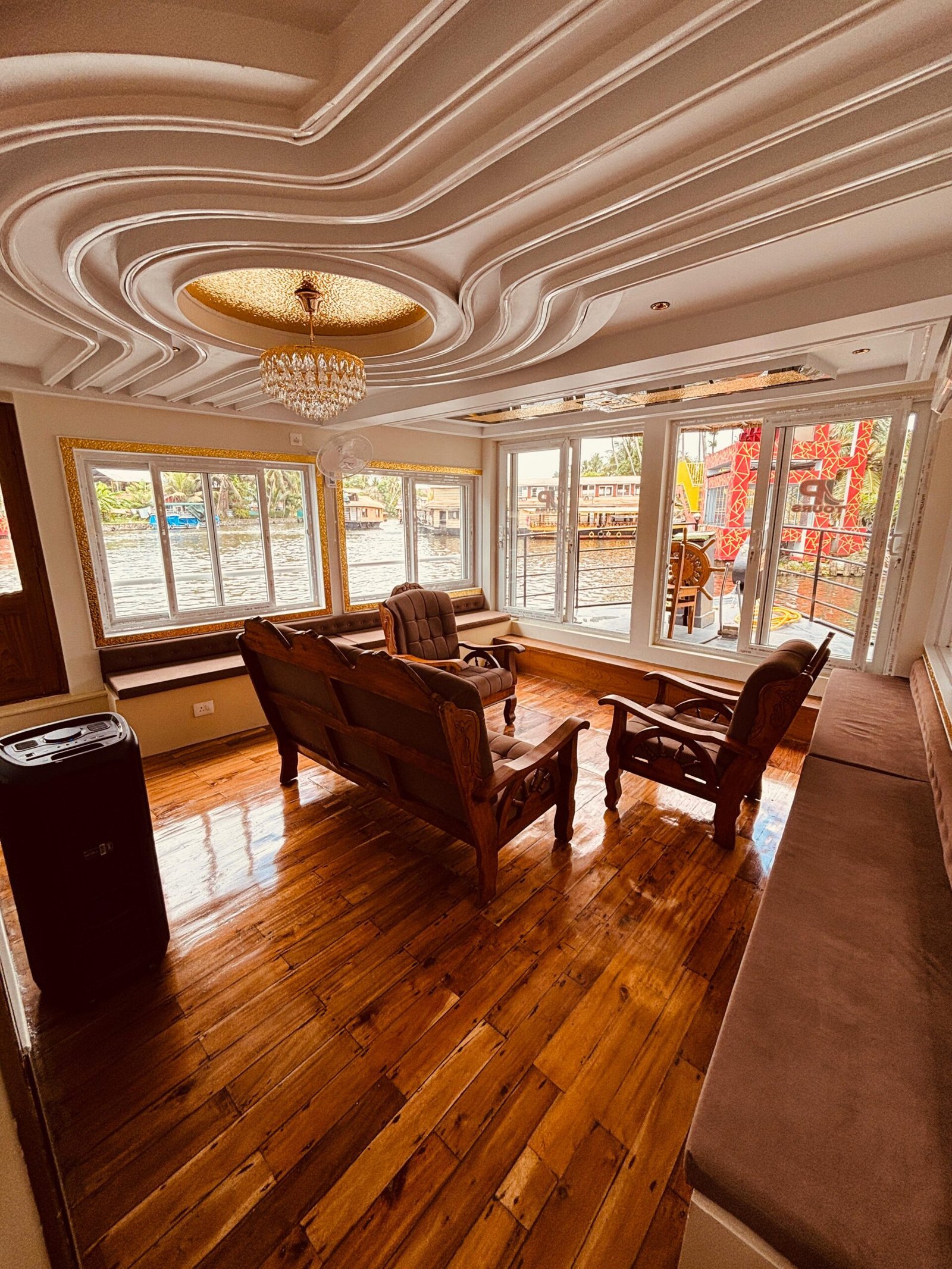 Spacious Living Area with Windows Spacious indoor living area with panoramic windows, wooden seating, and a sound system on a premium Kerala houseboat.