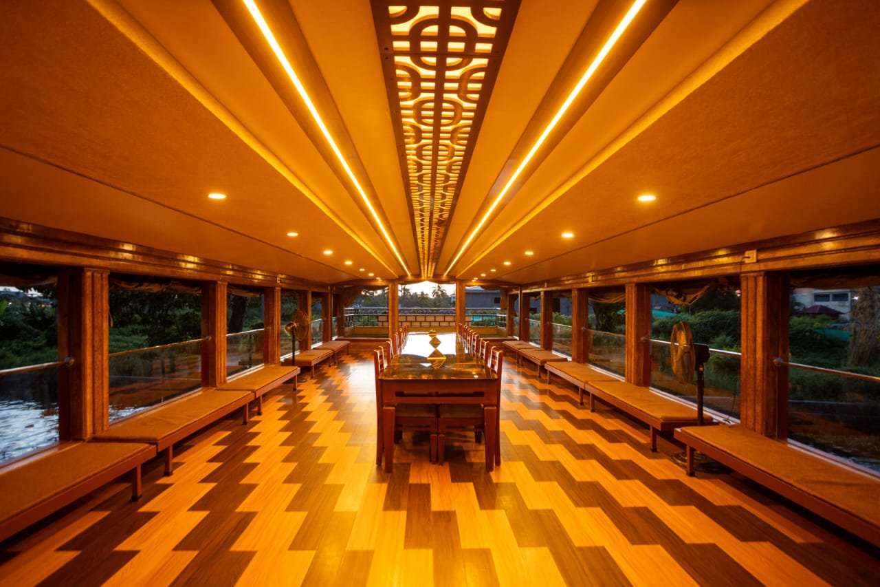 Spacious Lounge & Dining - 6 Bedroom Houseboat Alleppey Expansive lounge and dining hall with beautiful wooden flooring and ceiling lights inside a 6 bedroom houseboat alleppey.