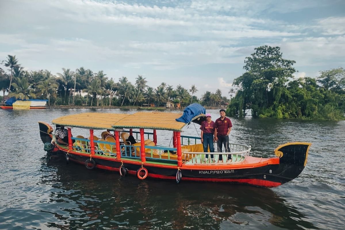 Two people standing at the bow of a colorful traditional boat with a woven roof during a scenic shikara ride in Alleppey backwaters.